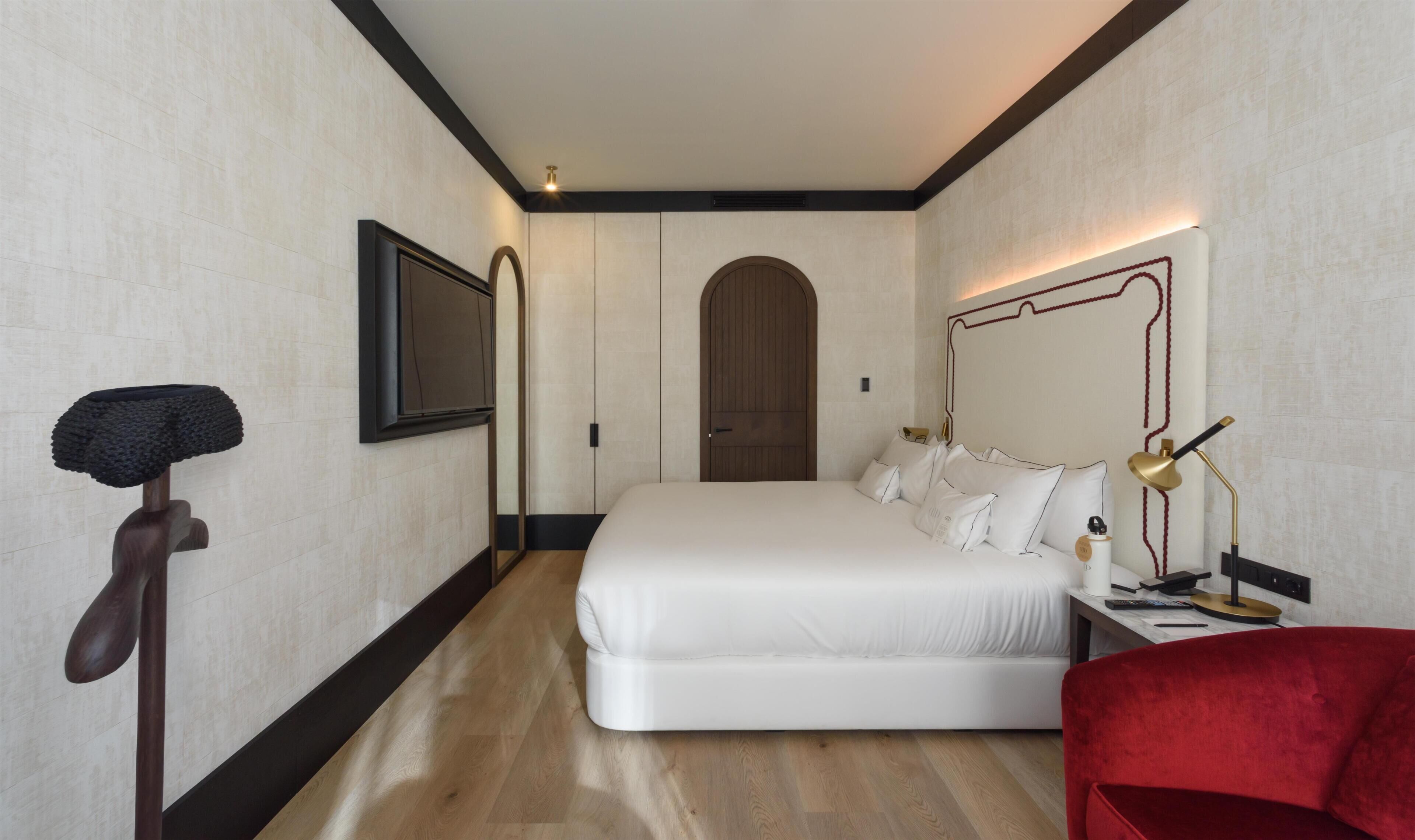 A hotel room with a large bed, a wooden floor, a red chair, and a desk with a lamp.