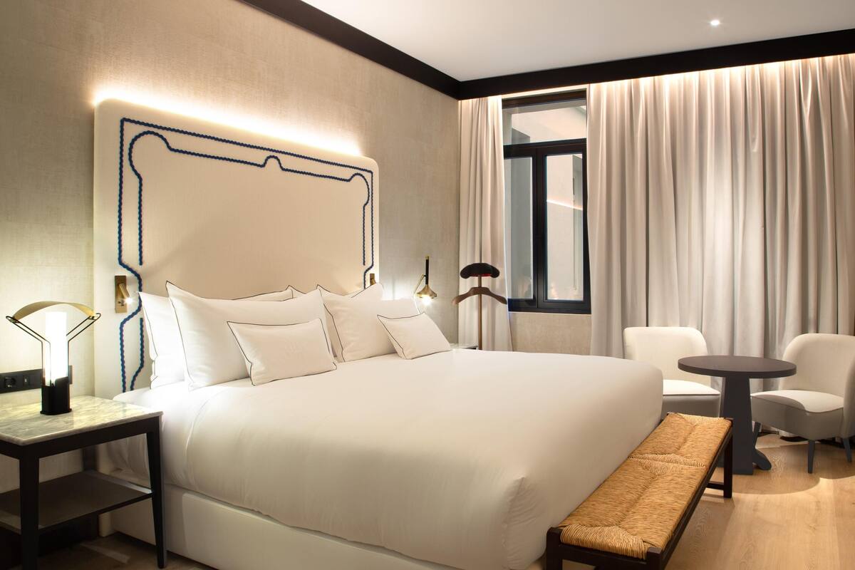 A hotel room with a large bed, a bedside table, a bench, two chairs, and a window with curtains.