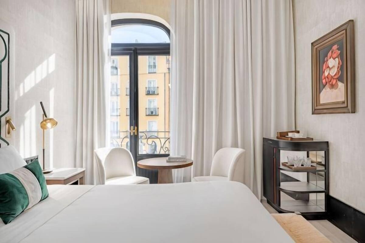 A hotel room with a bed, a small table, two chairs, a round table, a mirror, and a view of buildings through a door.