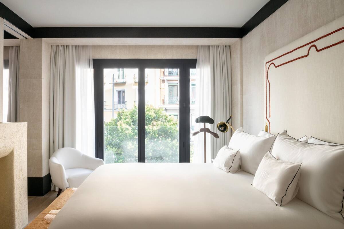 A hotel room with a large bed, a chair, a floor lamp, and a view of buildings through a large window.