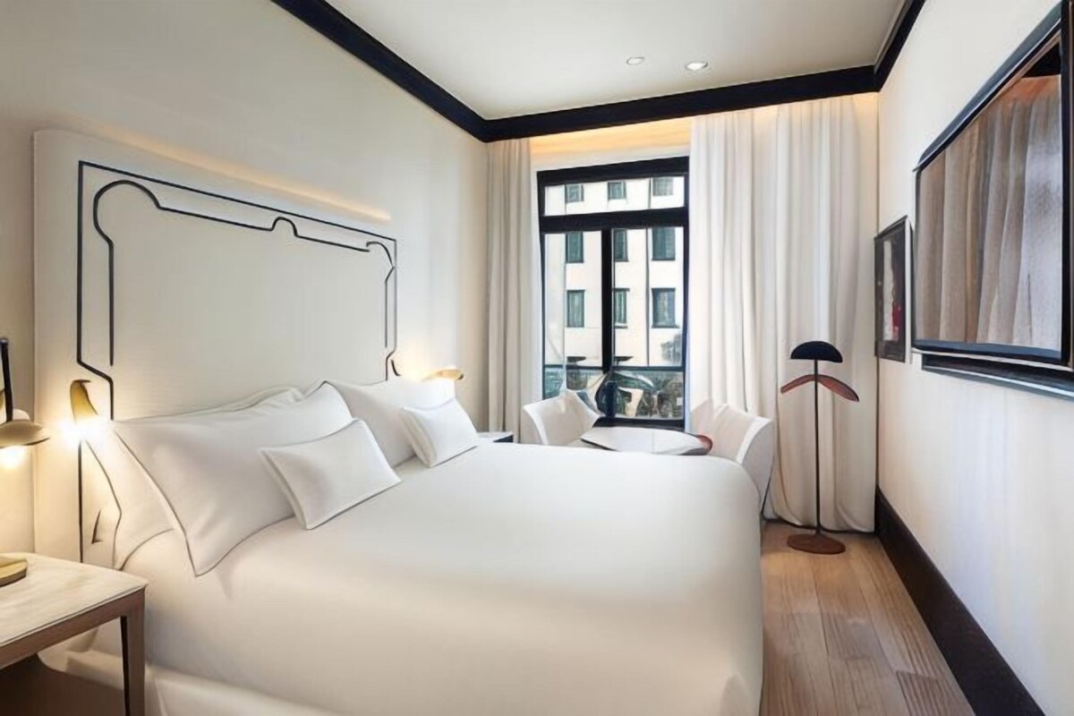 A hotel room with a large bed, bedside tables, a desk, a chair, a floor lamp, and a window with curtains.