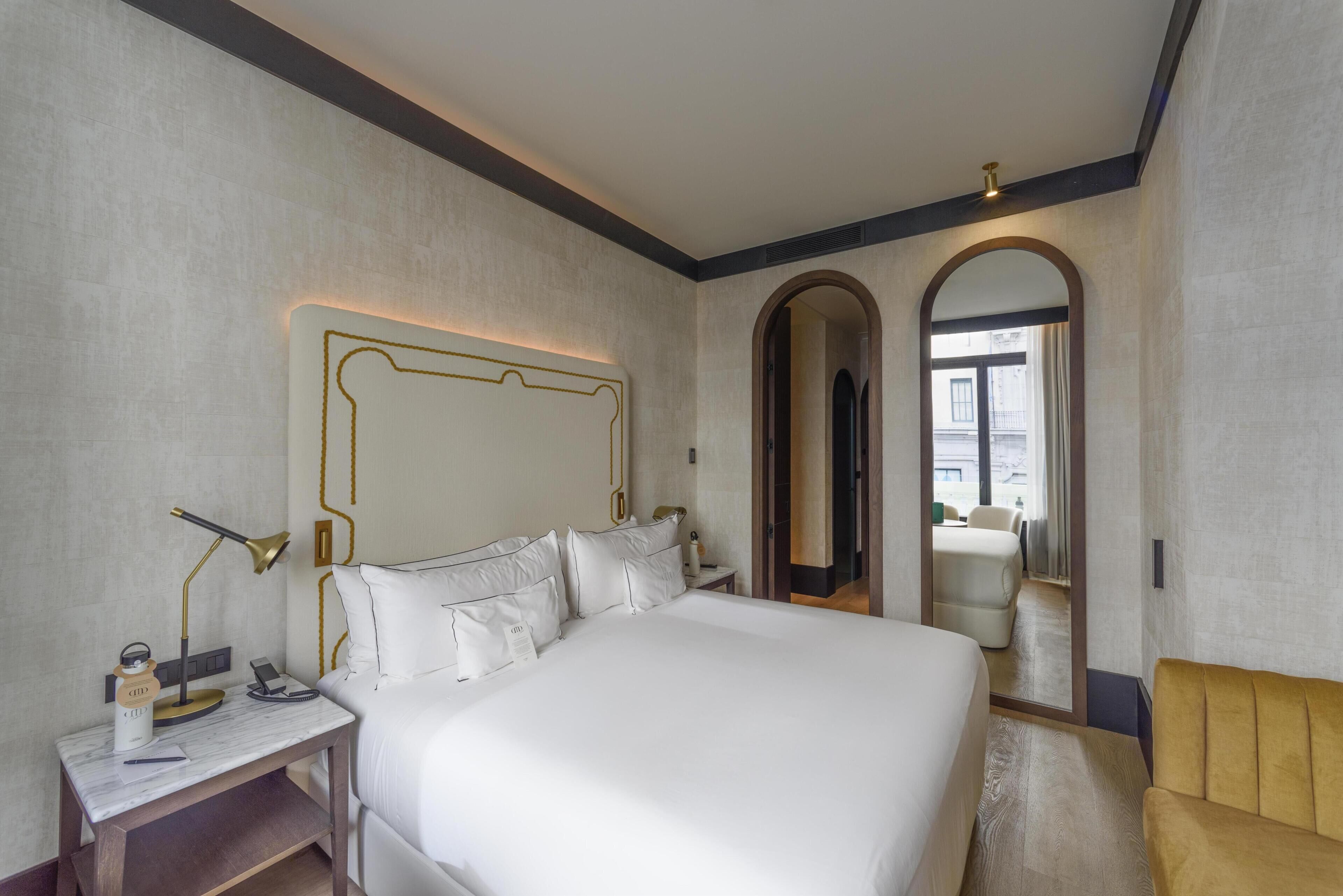 Una habitación de hotel con una cama grande, mesita de noche, lámpara y un espejo.