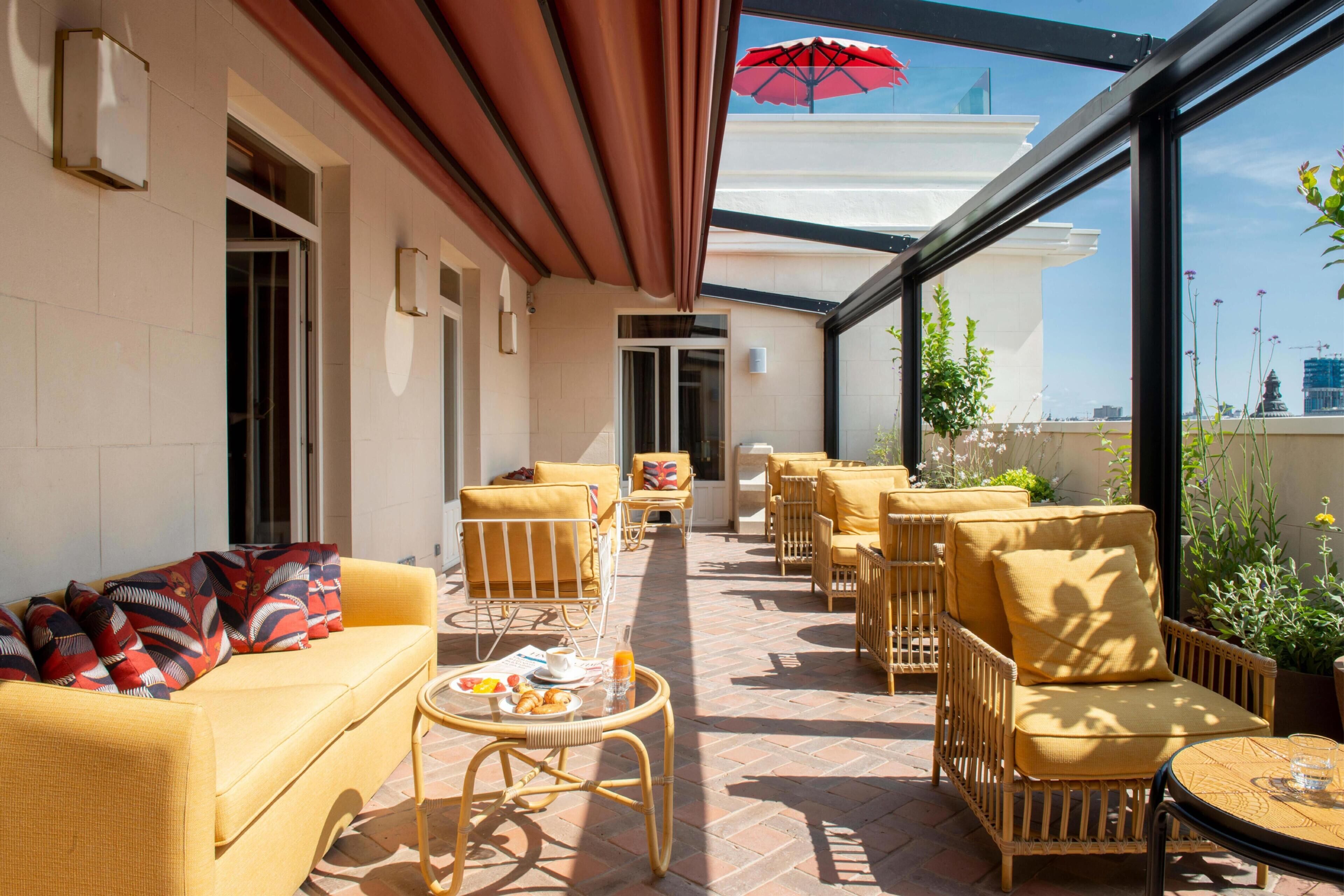 A sunlit outdoor seating area with yellow sofas and wicker chairs, a glass-enclosed patio, and a view of the cityscape.