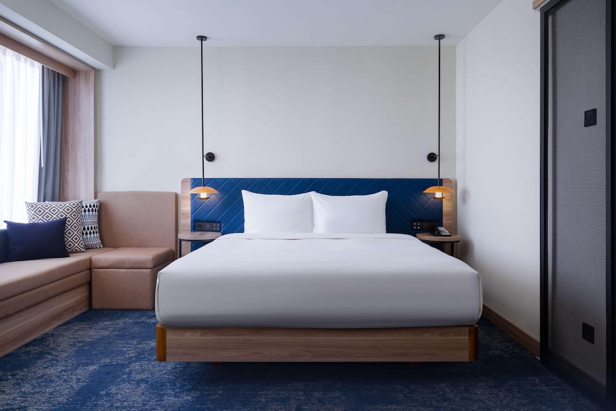 A hotel room with a bed, a sofa, two bedside lamps, a nightstand, and a window with curtains.