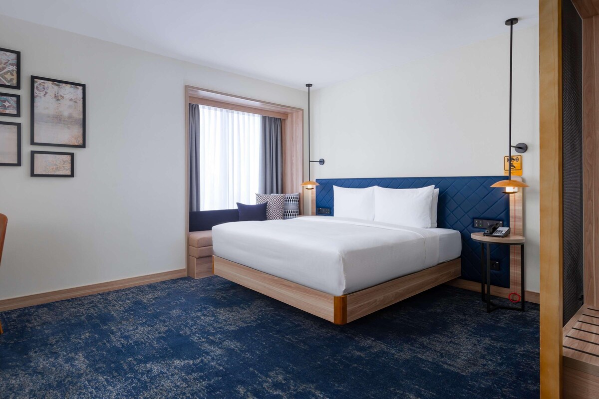 Een hotelkamer met een bed, een stoel, een bureau, een lamp en kunstwerken in een lijst aan de muur.