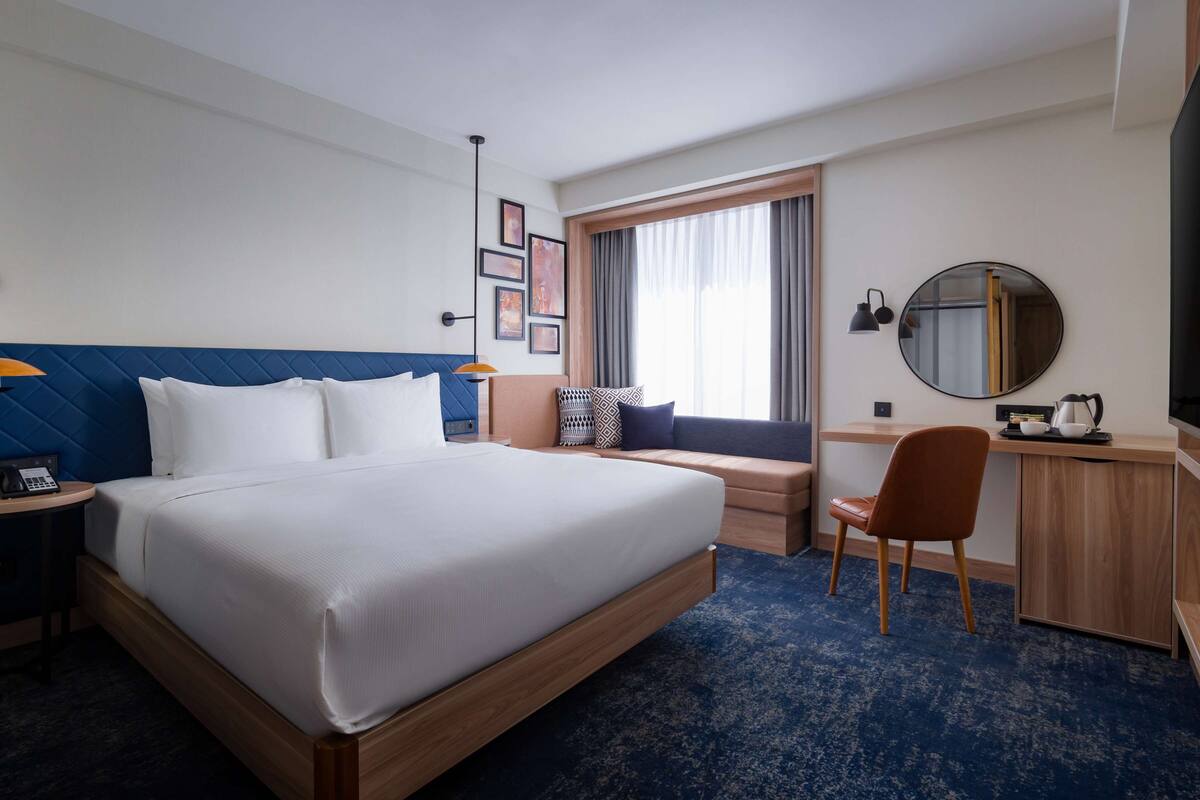 A hotel room with a large bed, a desk, a chair, a round mirror, and a window with curtains.