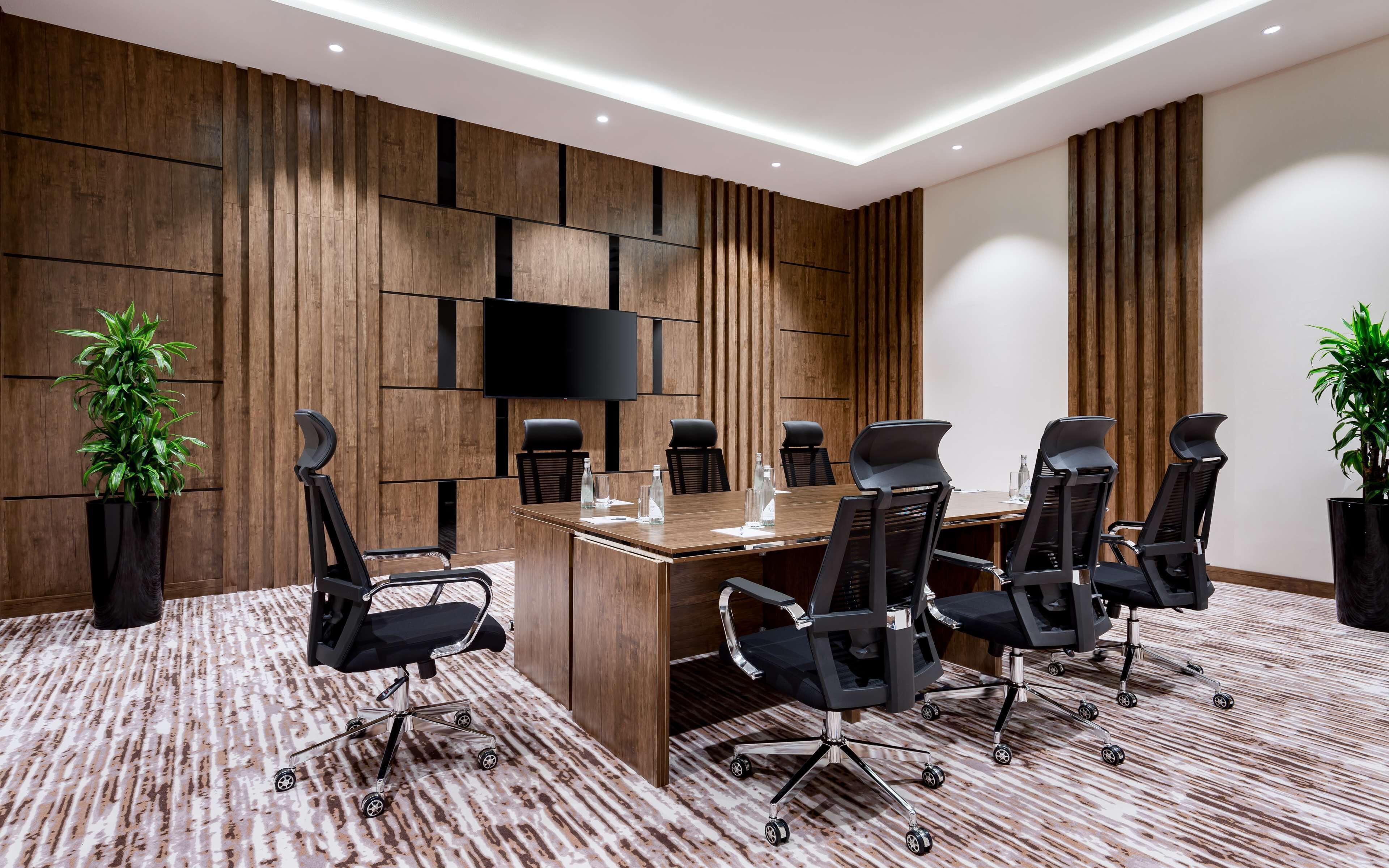 A conference room with a long table, black chairs, a flat-screen TV, and wooden panel walls.