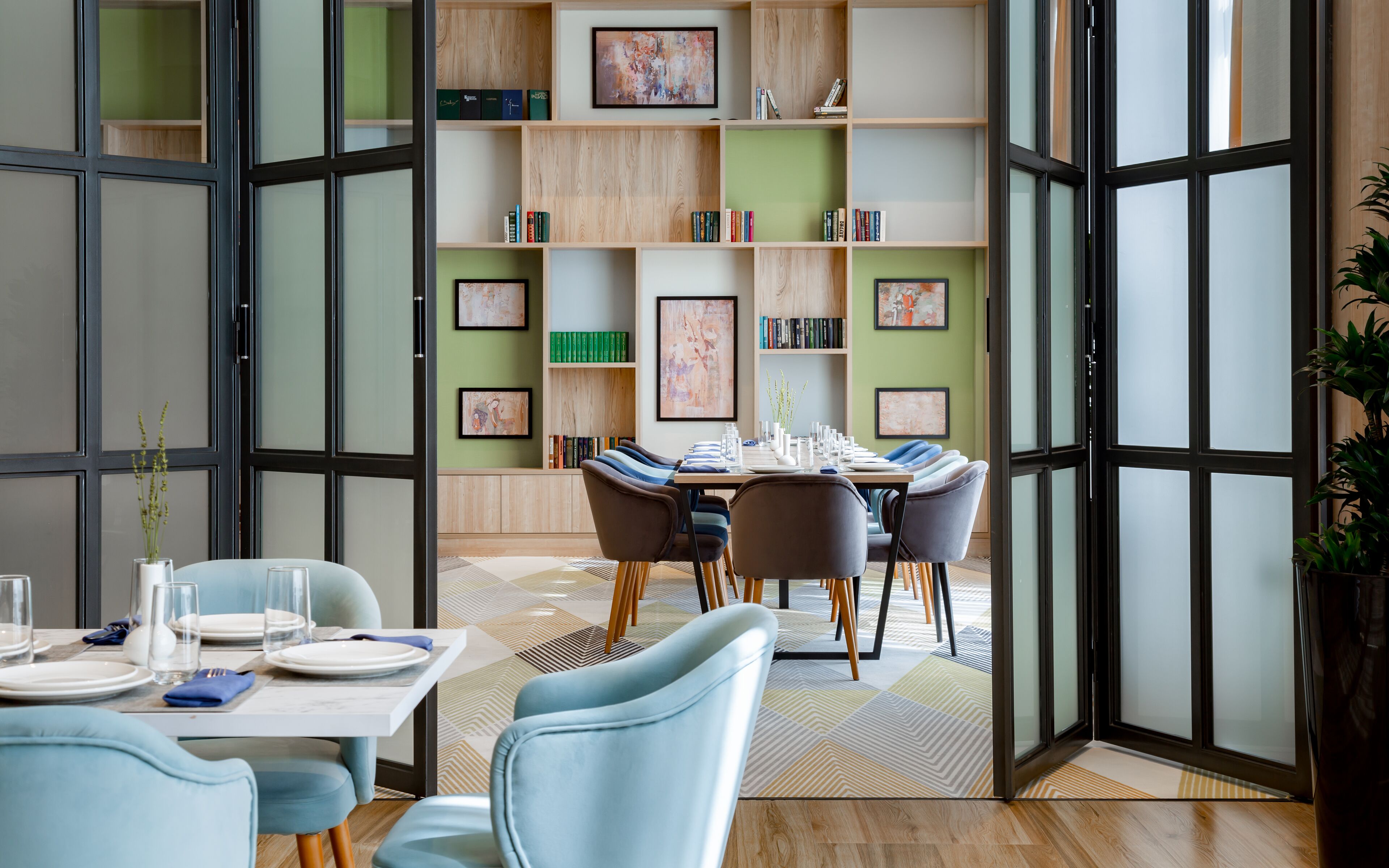 A modern dining area with a table set for four, surrounded by chairs. A bookshelf with various books and framed artwork is visible in the background.