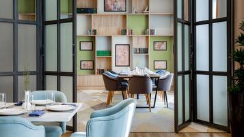 A modern dining area with a table set for four, surrounded by chairs. A bookshelf with various books and framed artwork is visible in the background.