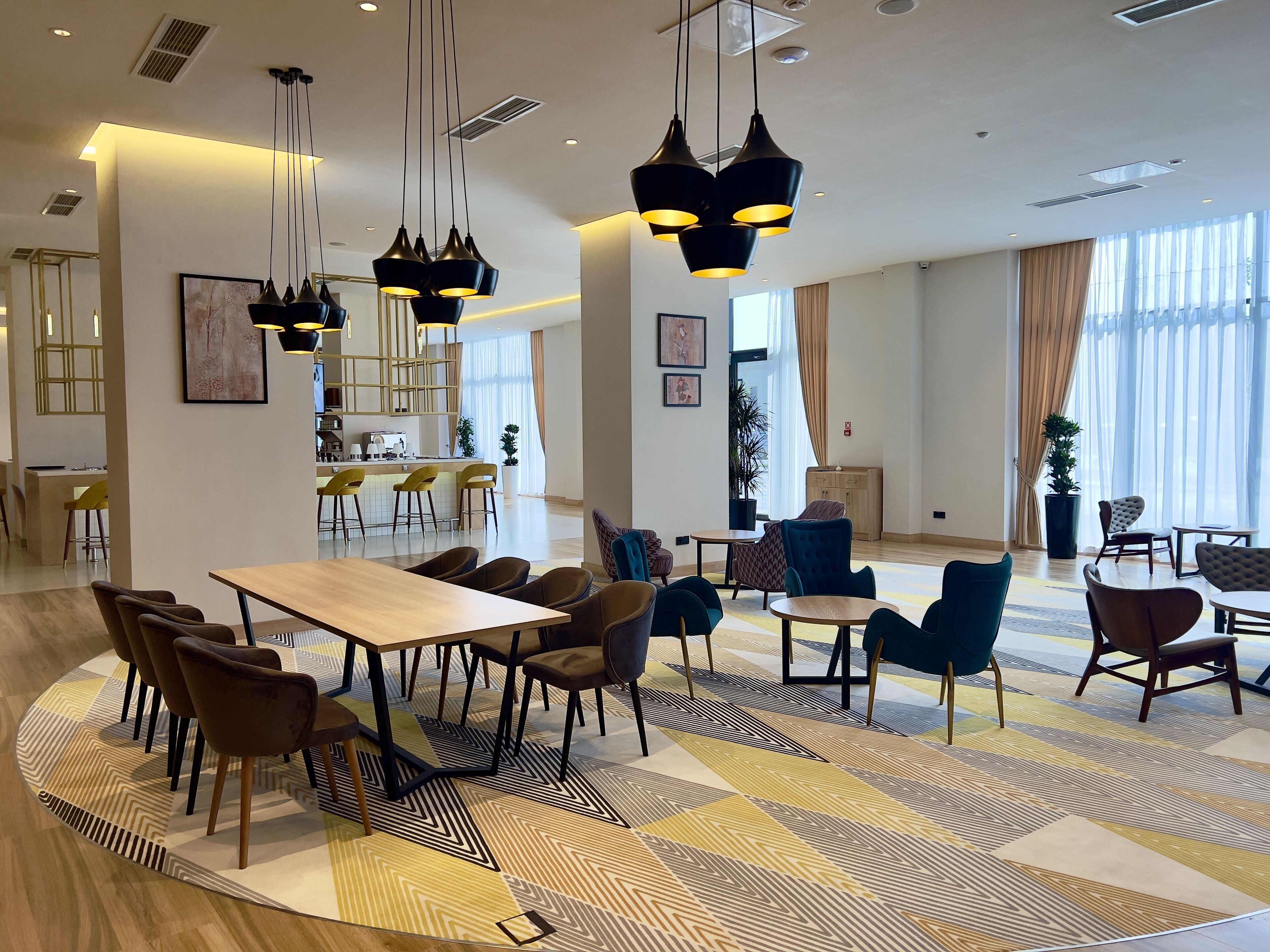 A modern hotel lobby with a central dining area, multiple seating options, and large windows providing natural light.