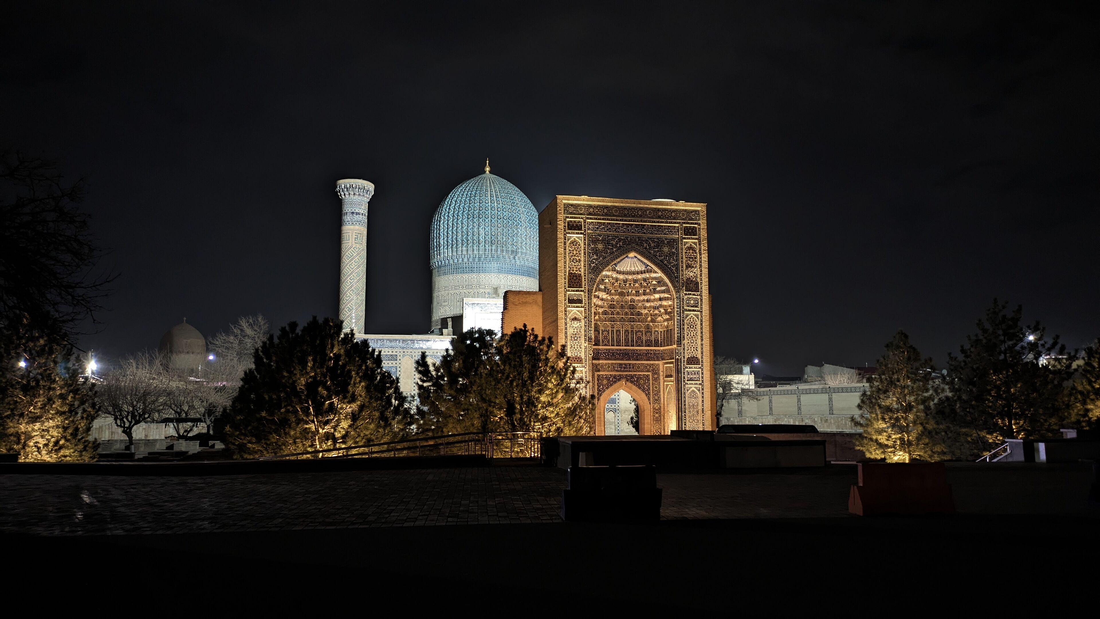 Una vista nocturna de un edificio histórico con una cúpula grande y un minarete, rodeado de árboles y una zona pavimentada.