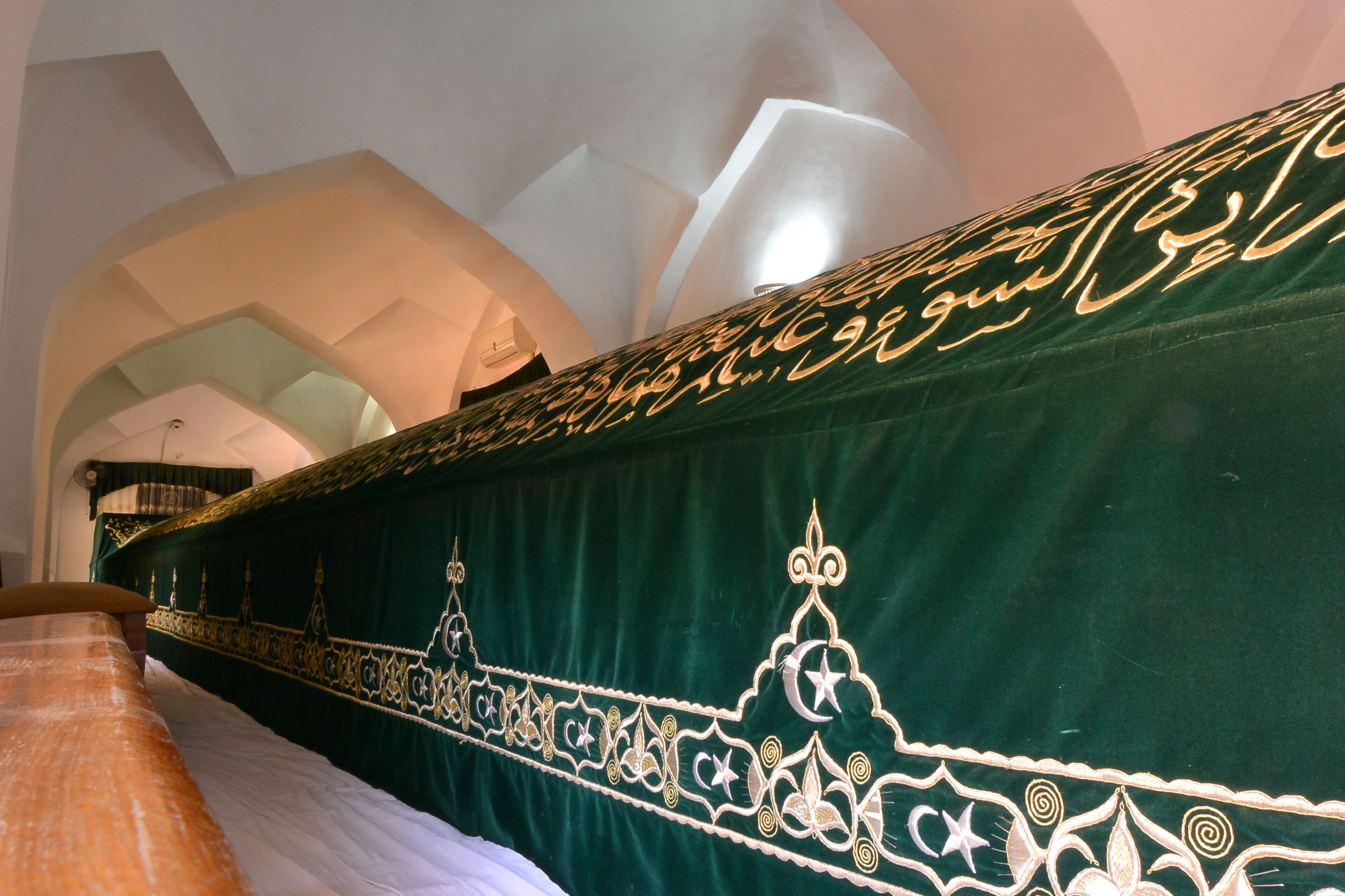 Ein Zimmer mit einem dunkelgrünen, gepolsterten Kopfteil mit aufwendiger goldener Stickerei, einem hölzernen Bettgestell und einer weißen Bettdecke.