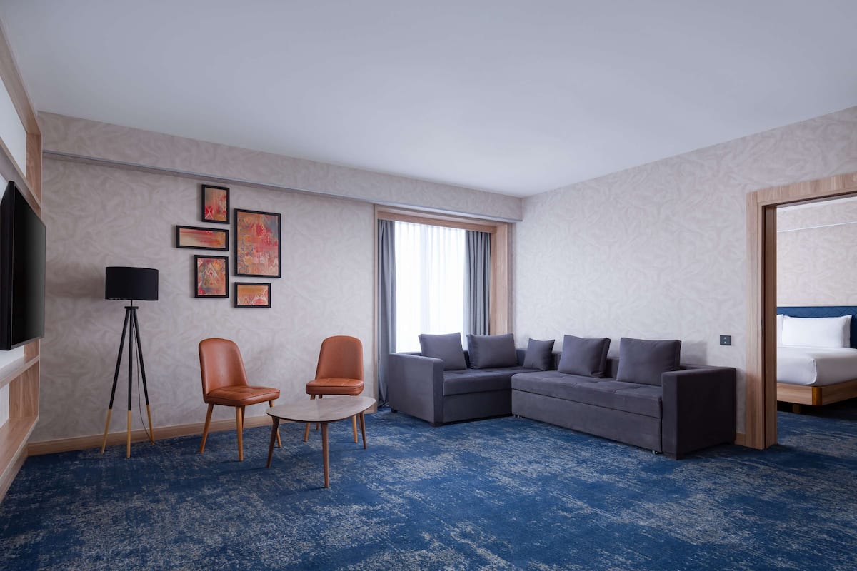 A hotel room with a large gray sectional sofa, two armchairs, a small round table, a flat-screen TV mounted on a wooden cabinet, and a large window with curtains.