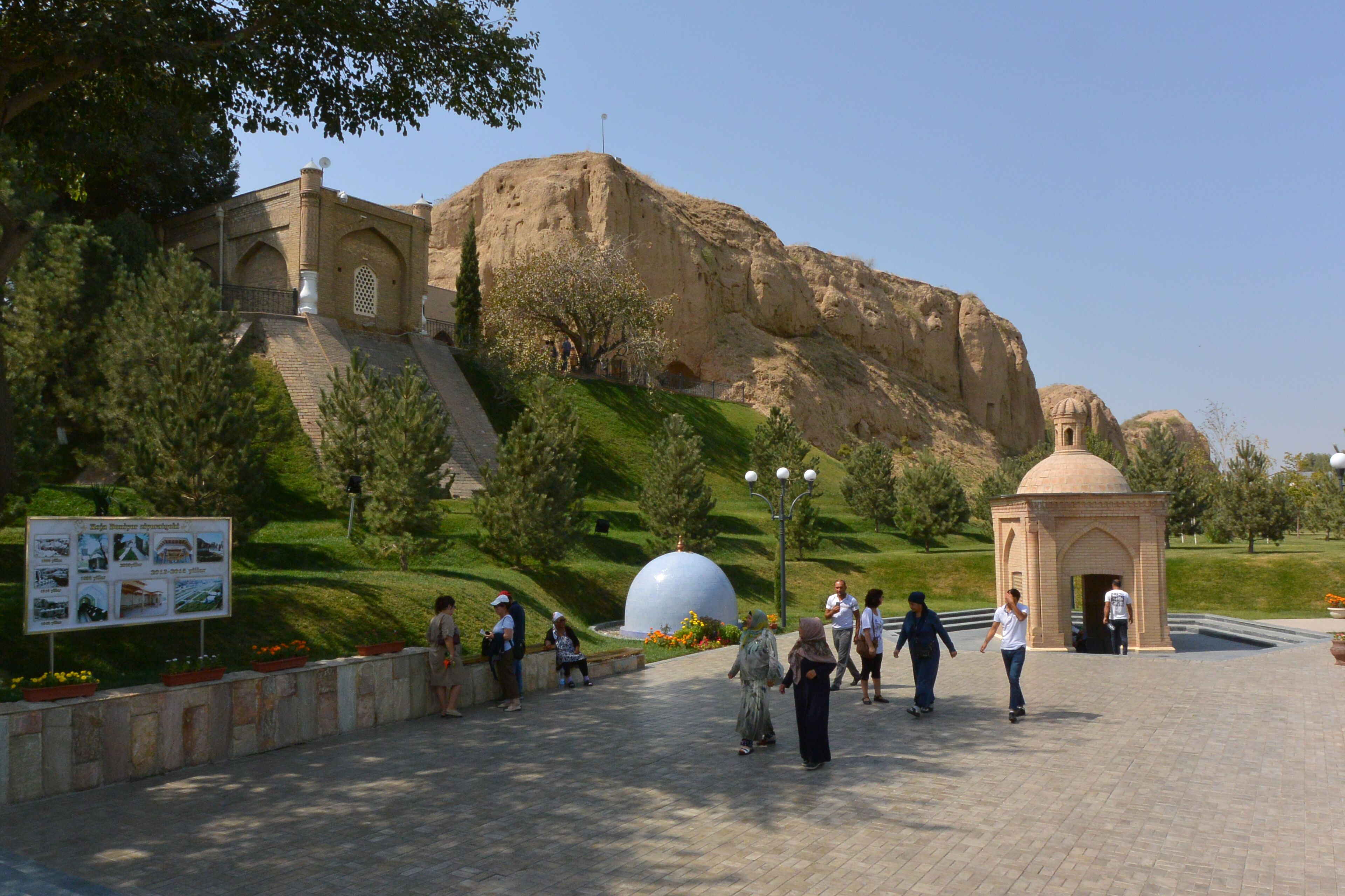A paved walkway with a large dome structure, a small building with a dome, and a hill with a large rock formation in the background.