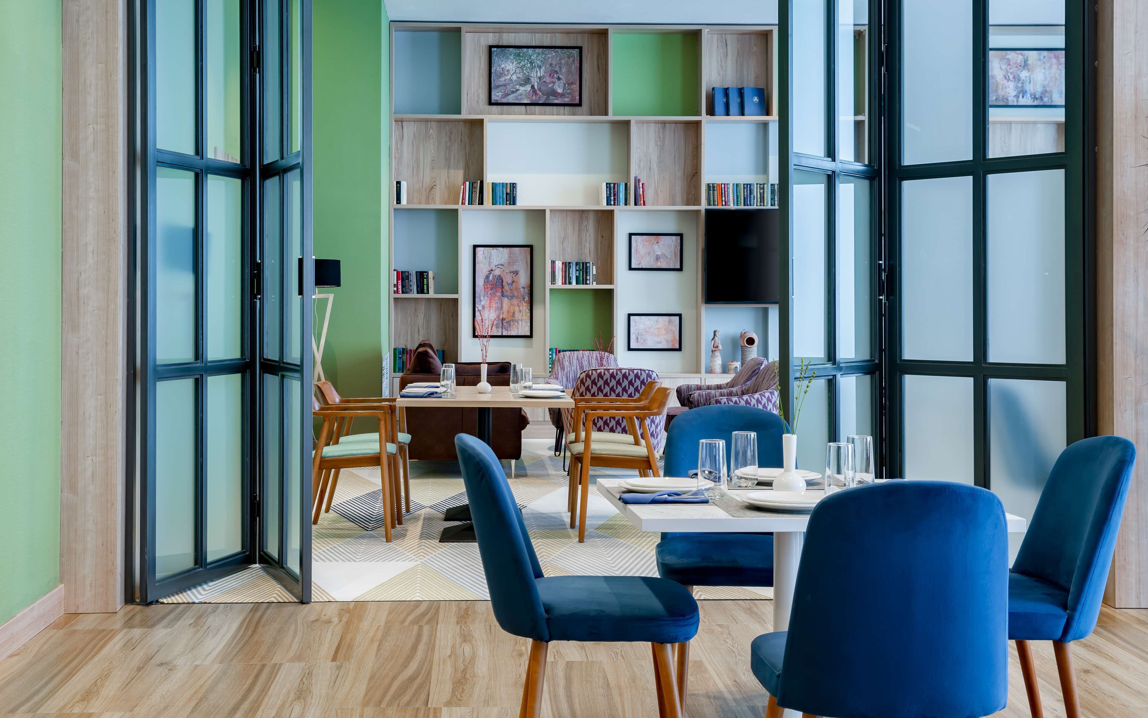 A modern dining area with a table set for four, surrounded by blue chairs, and a bookshelf filled with books and framed art in the background.