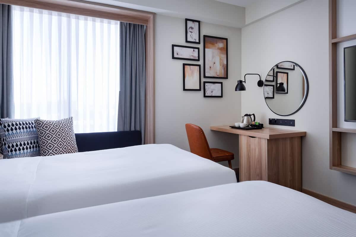 A hotel room with two beds, a desk, a chair, a mirror, and artwork on the wall.