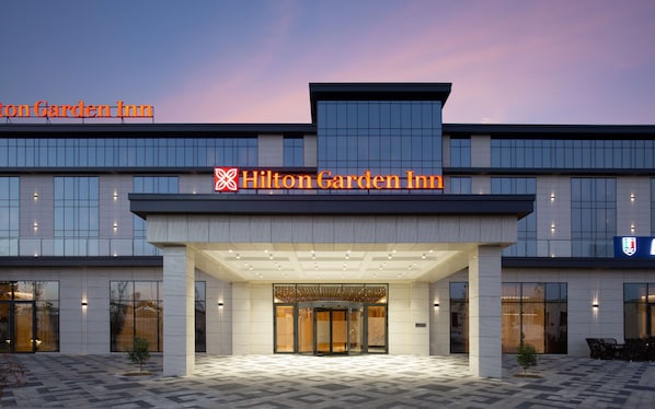 A modern hotel entrance with a glass facade, a prominent sign for Hilton Garden Inn, and a covered entrance area. - Hilton Garden Inn Samarkand (Samarkand)
