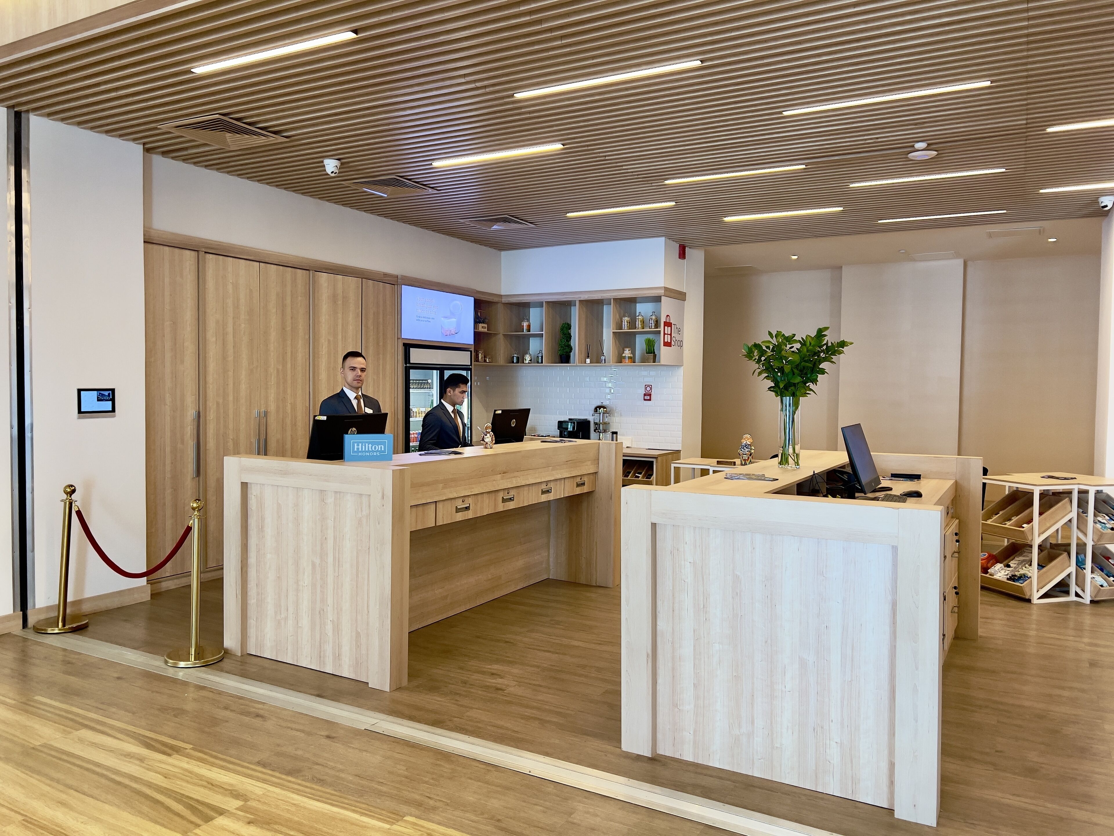 Un hall d’accueil d’hôtel doté d’un plancher en bois, d’un design de plafond moderne et d’un comptoir de réception où travaillent deux employés.