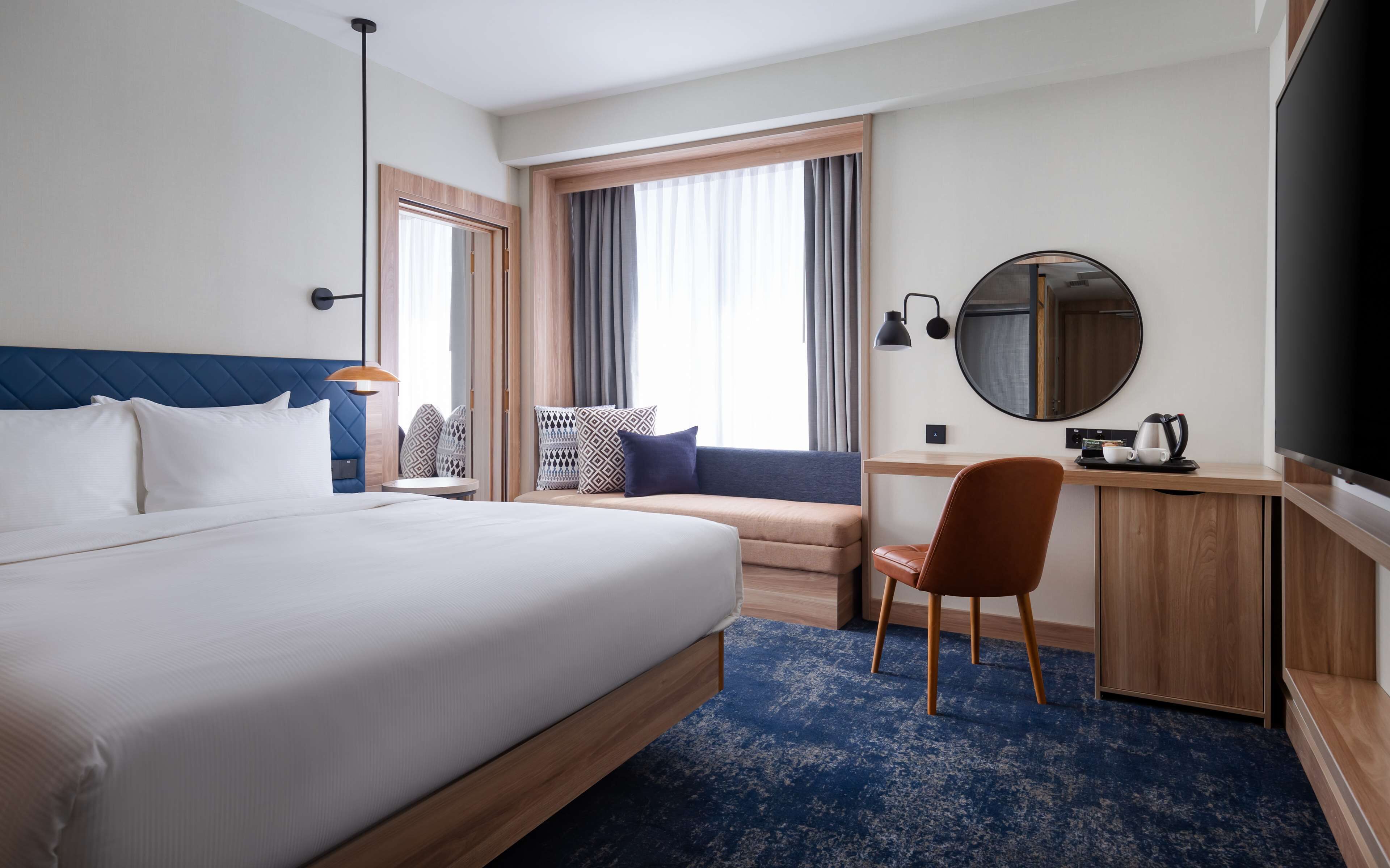 A hotel room with a large bed, a desk, a chair, a round mirror, and a television.