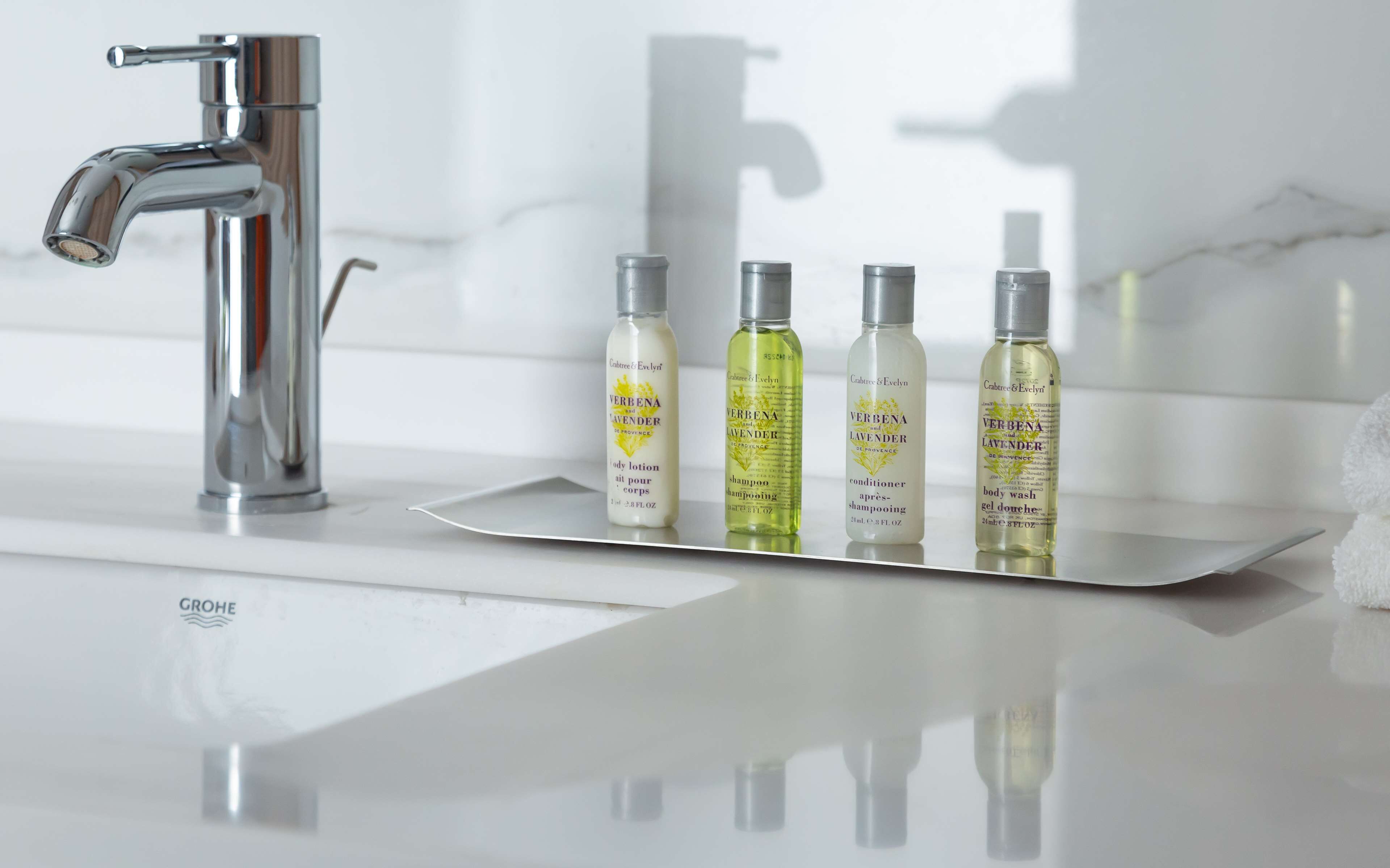 Un baño con un grifo cromado, un lavamanos blanco y una bandeja con cuatro botellas de Crabtree & Evelyn Verbenas.