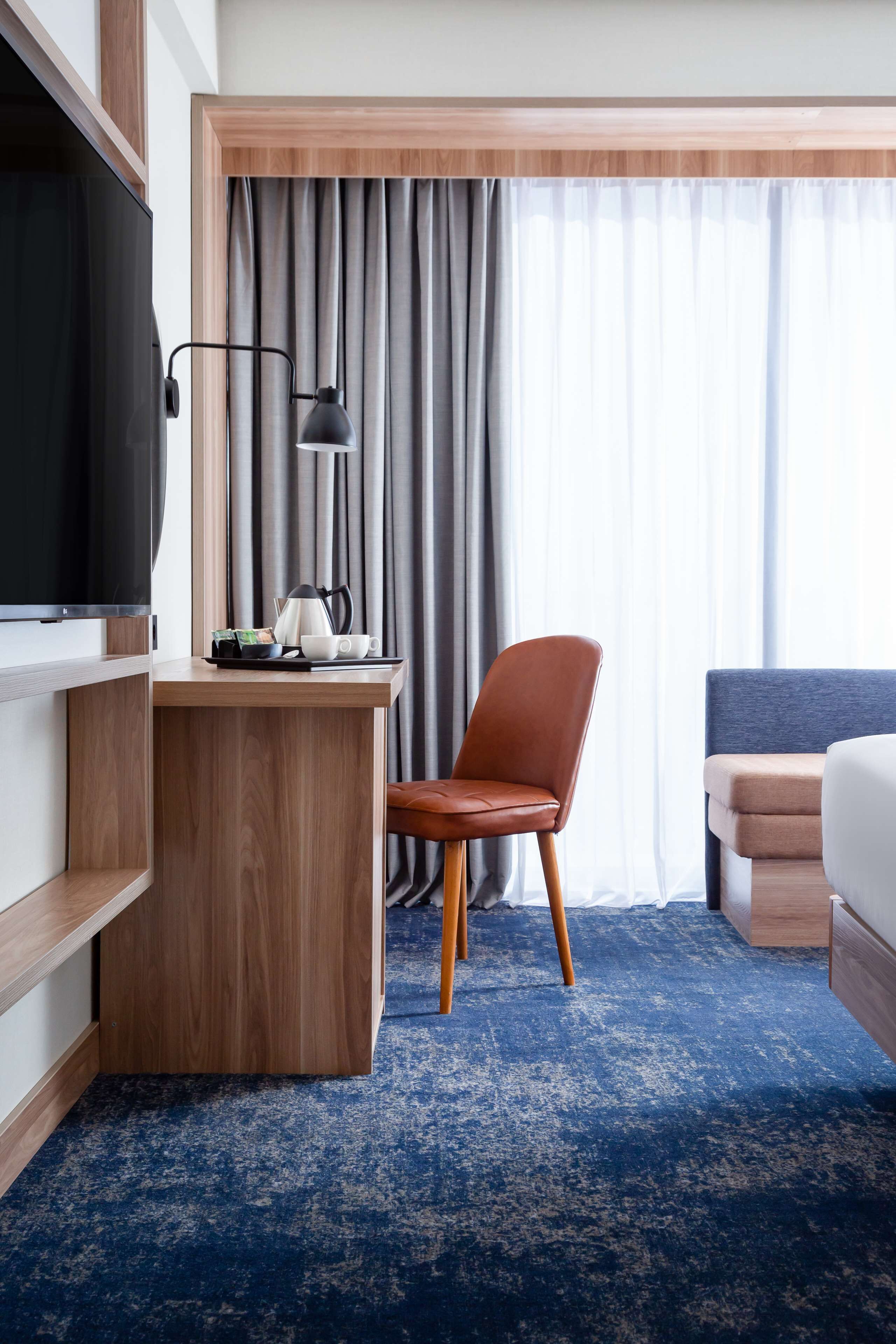 A hotel room with a wooden desk, a television, a chair, a bed, and a large window with curtains.