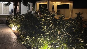 A well-lit outdoor area with a covered seating area, surrounded by lush greenery and a paved walkway. - Villa Samadoel (Santa Maria del Focallo)