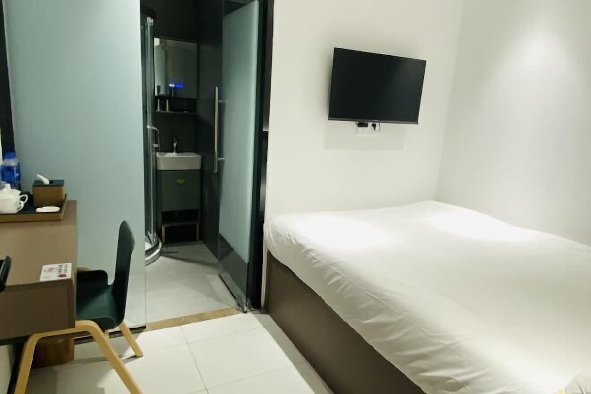 A hotel room with a bed, a desk with a chair, a TV mounted on the wall, and a bathroom visible through a glass door.