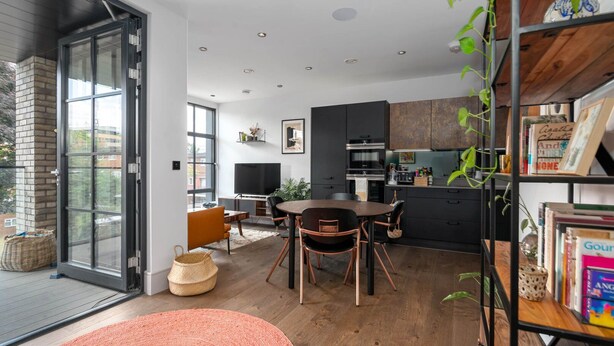Een moderne keuken met donkere kastjes, een eethoek met een ronde tafel en stoelen, en een groot raam dat natuurlijk licht binnenlaat.