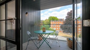 Een balkon met een glazen tafel en twee stoelen, met uitzicht op een woongebied met bakstenen gebouwen en bomen.
