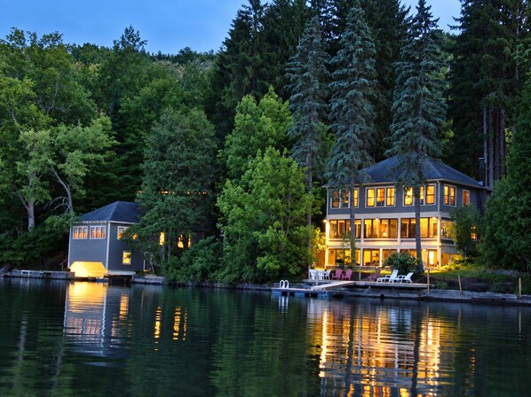 A two-story house with a dock and chairs by the water, surrounded by trees.