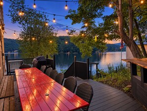 A wooden deck with a red table and wicker chairs, overlooking a lake with trees and a sunset.