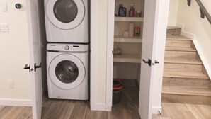 A laundry room with two stacked washing machines, a shelf with cleaning supplies, and a wooden staircase. - Lovely 2 Bedroom Guest Suite in Southwest Edmonton (Edmonton)