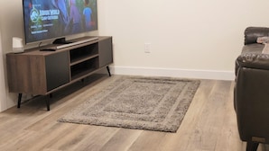 A living room with a flat-screen TV on a wooden stand, a brown leather sofa, and a beige rug. - Lovely 2 Bedroom Guest Suite in Southwest Edmonton (Edmonton)