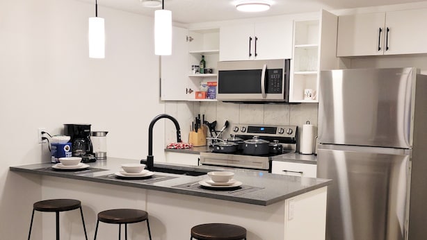 A modern kitchen with a stainless steel refrigerator, a microwave, and an oven. There are three stools at the island, a coffee maker, and a sink with a faucet. - Lovely 2 Bedroom Guest Suite in Southwest Edmonton (Edmonton)