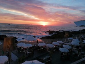 A beachside restaurant with outdoor seating and a view of the sunset over the ocean.