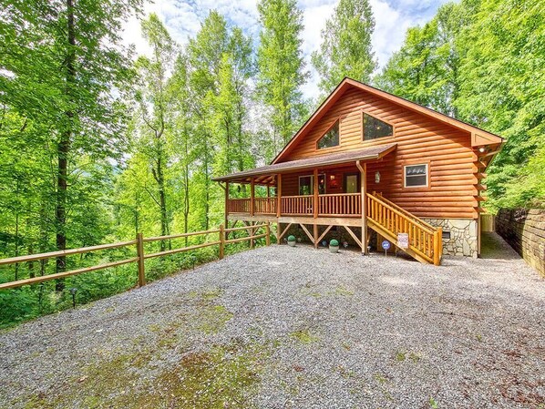 A log cabin with a porch and a stone foundation, surrounded by a gravel driveway and a wooden fence, set in a forest. - Log Cabin in a Gated Community! WIFI. Fire Pit w/ wood provided! Pet-friendly (Waynesville)