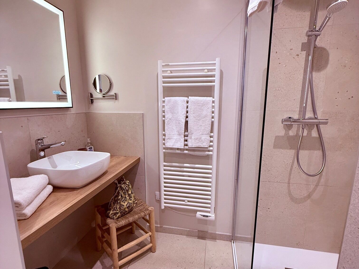 Ein Badezimmer mit Waschbecken, Spiegel, Handtuchhalter, Dusche und einem kleinen Hocker.