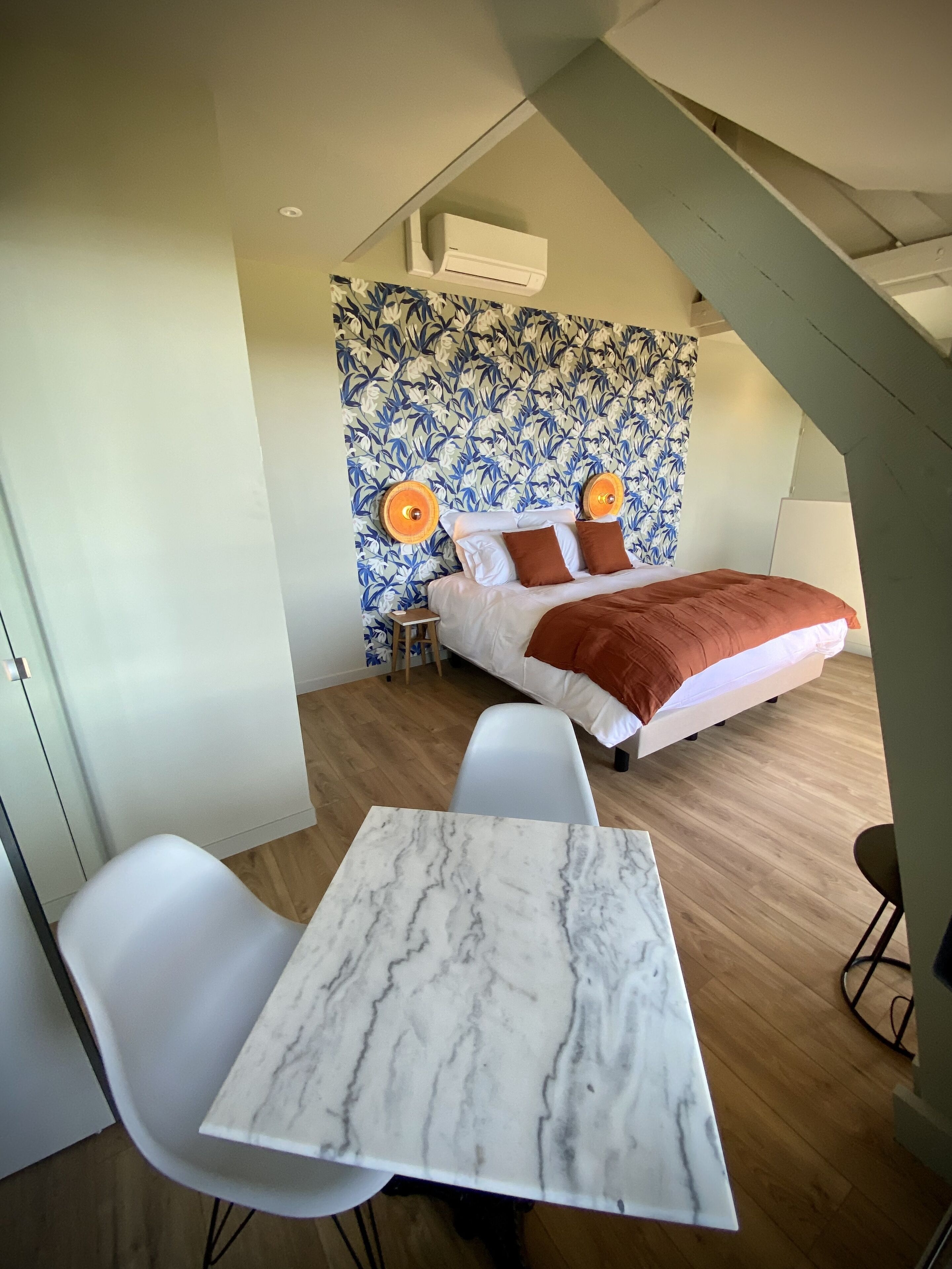A modern bedroom with a bed, a small table, and a chair. There is a patterned wall behind the bed and an air conditioning unit above it.