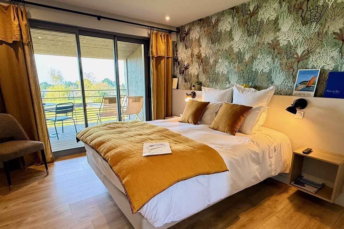 A bedroom with a large bed, a chair, a nightstand, and a view of the outdoors through a sliding door.