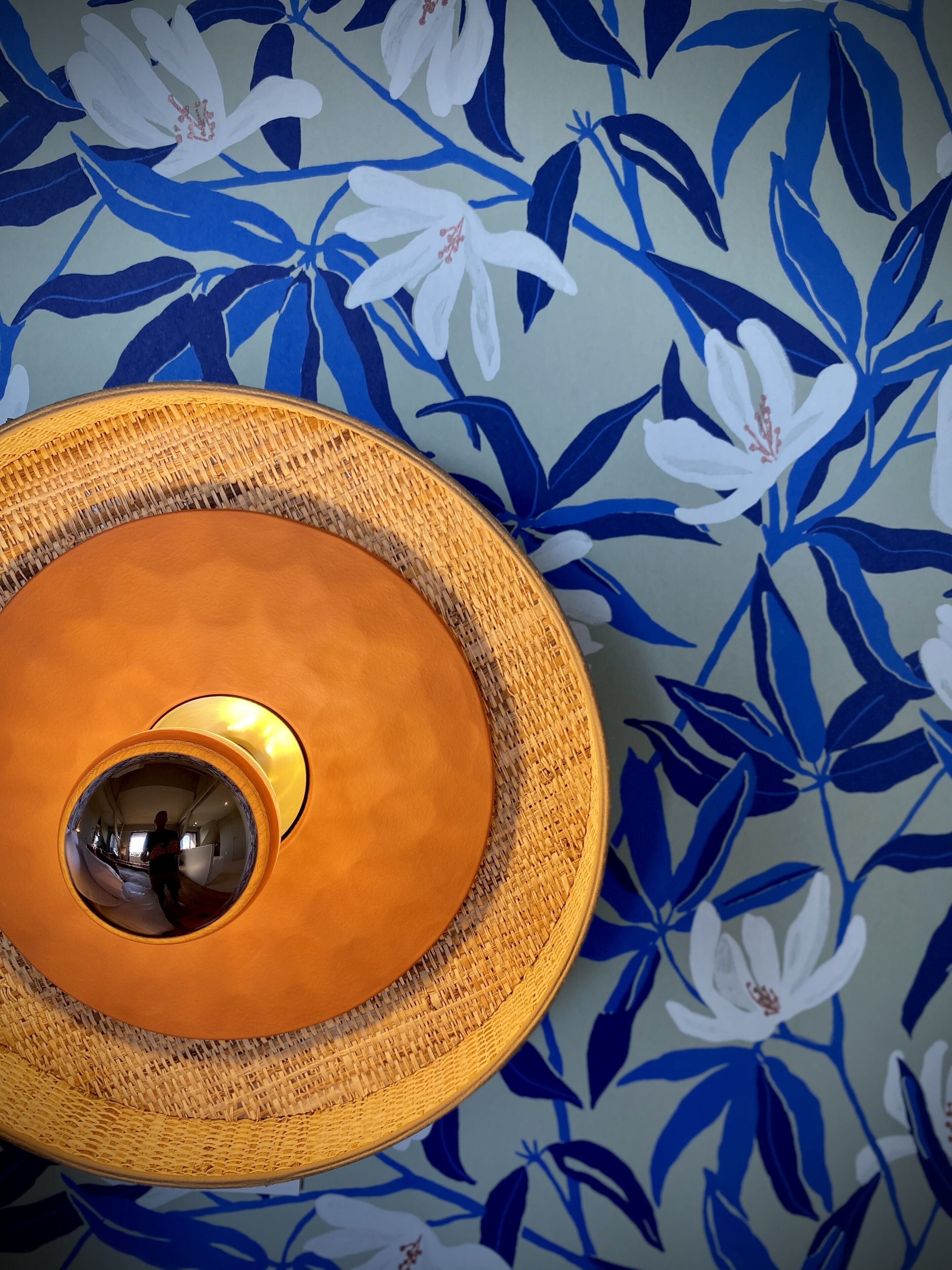 A circular, woven light fixture with a central reflective element, set against a wall with a floral pattern.