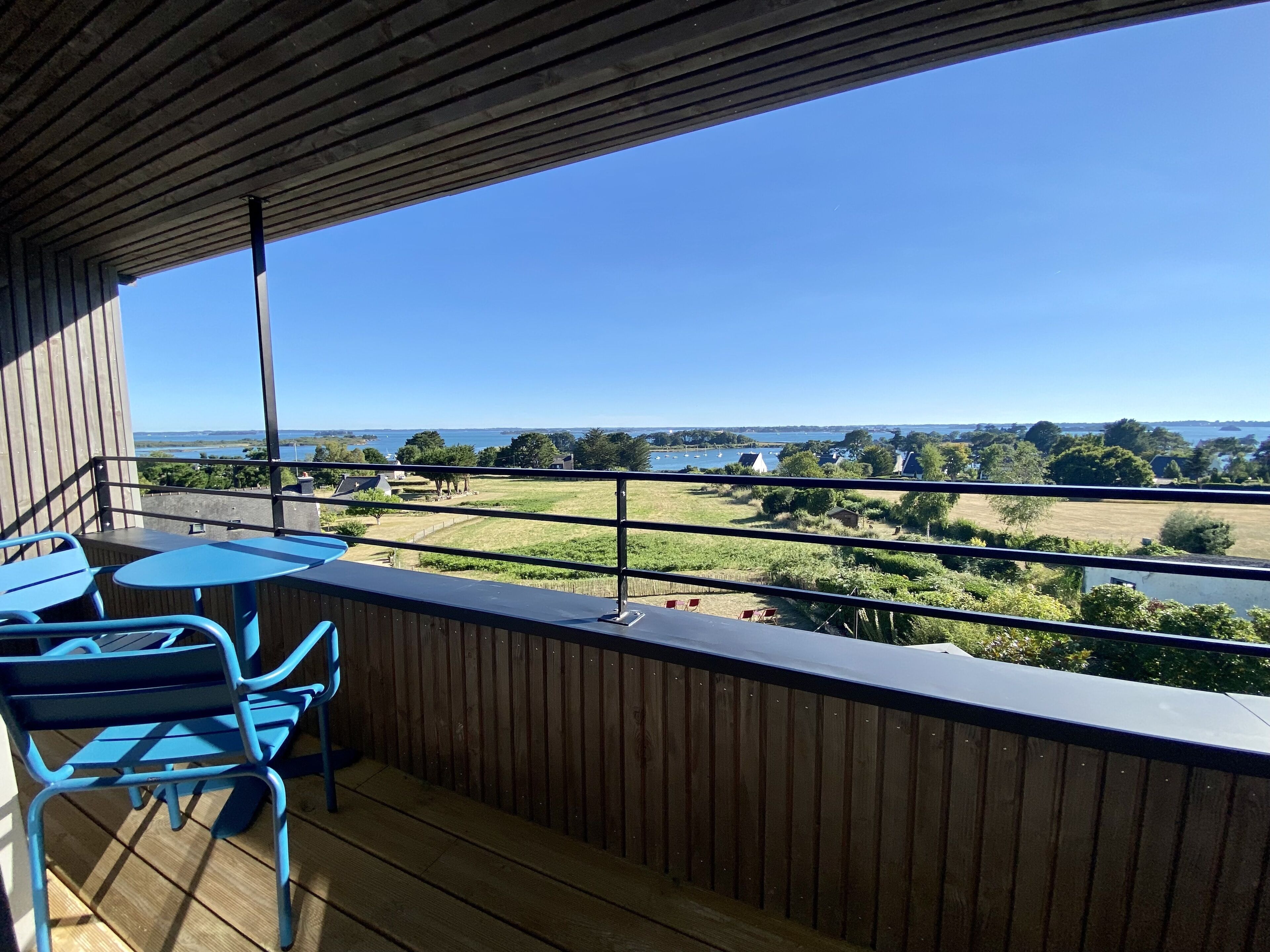 A balcony with a wooden floor and blue metal chairs, overlooking a grassy area and a body of water with boats.