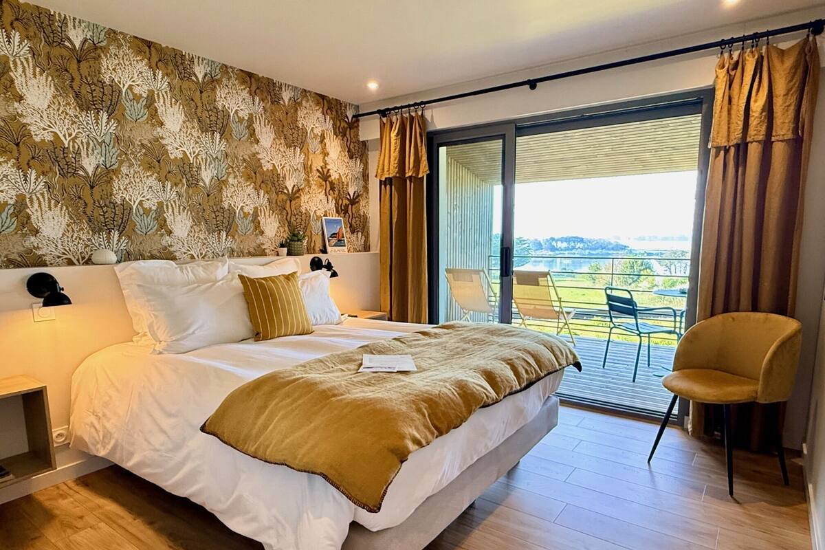 A bedroom with a large bed, a nightstand, a chair, a balcony with a view, and a patterned wall.