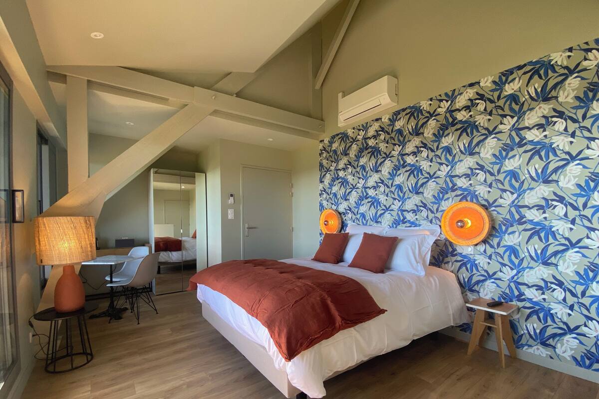 A modern hotel room with a large bed, a desk, and a chair. There is a mirror, a lamp, and a wall with a floral pattern.