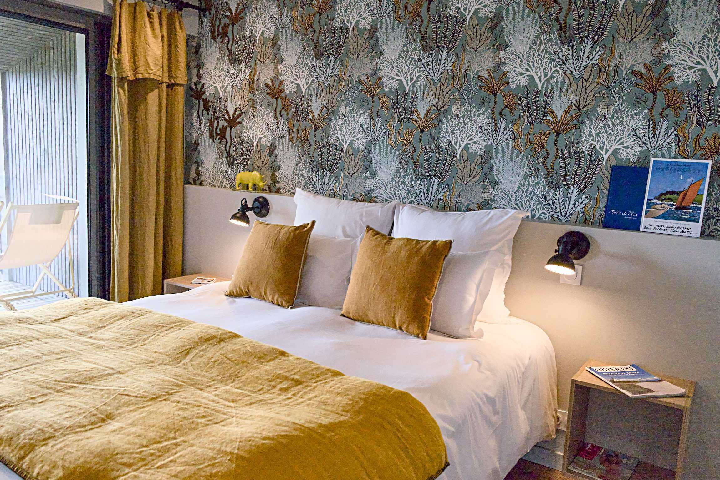 A hotel room with a bed, a nightstand, a chair, a window with curtains, and a wall with a leaf pattern.