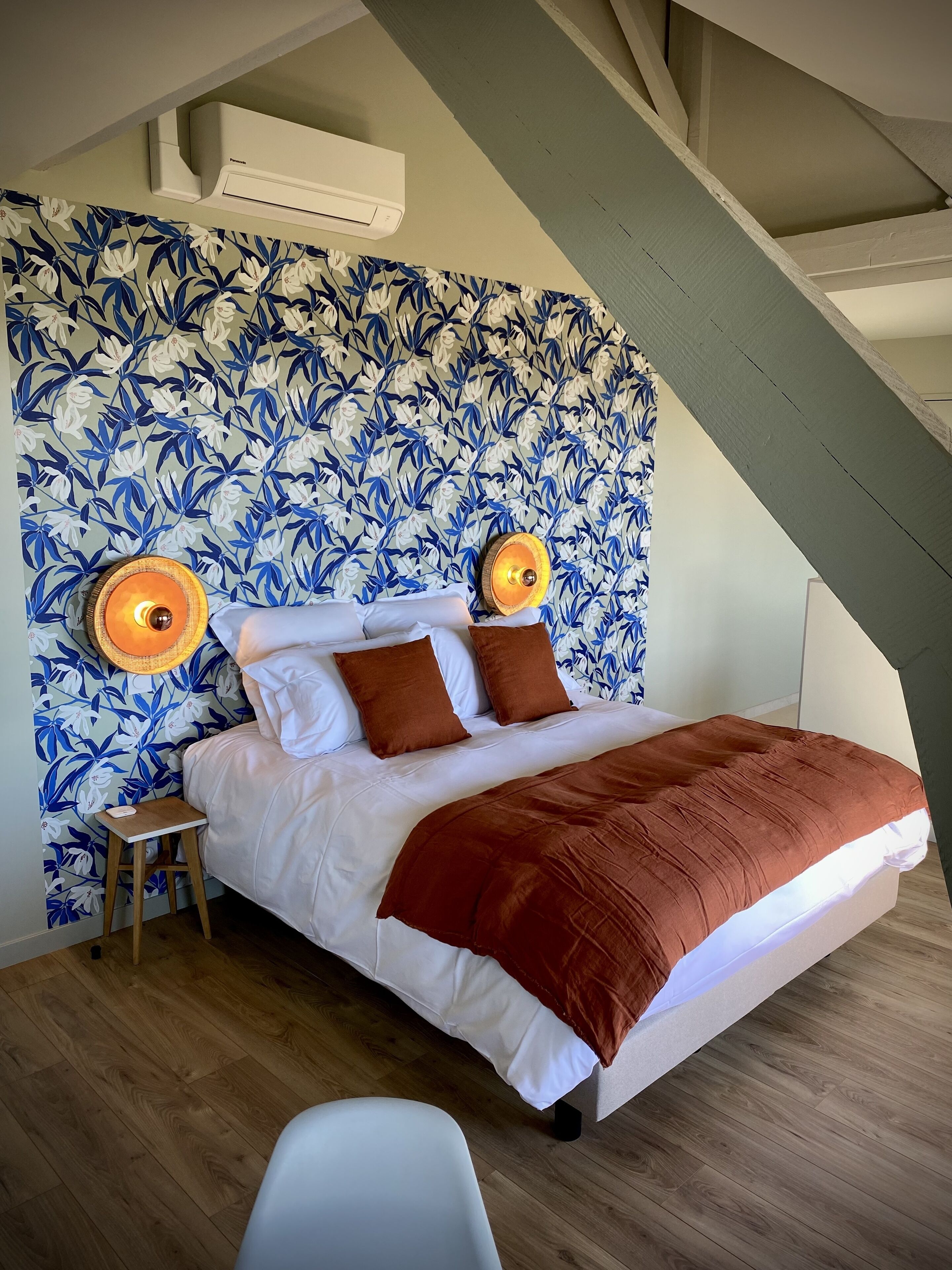 Ein Schlafzimmer mit blumigem Wandpapier, einem Bett mit weißer Bettwäsche und braunen Kissen, einem kleinen Holzschemel und einem weißen Stuhl.