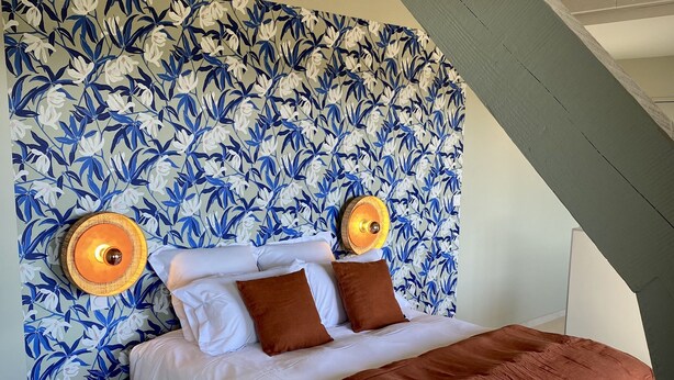 A bedroom with a floral wallpaper, a bed with white bedding and brown pillows, a small wooden stool, and a white chair.