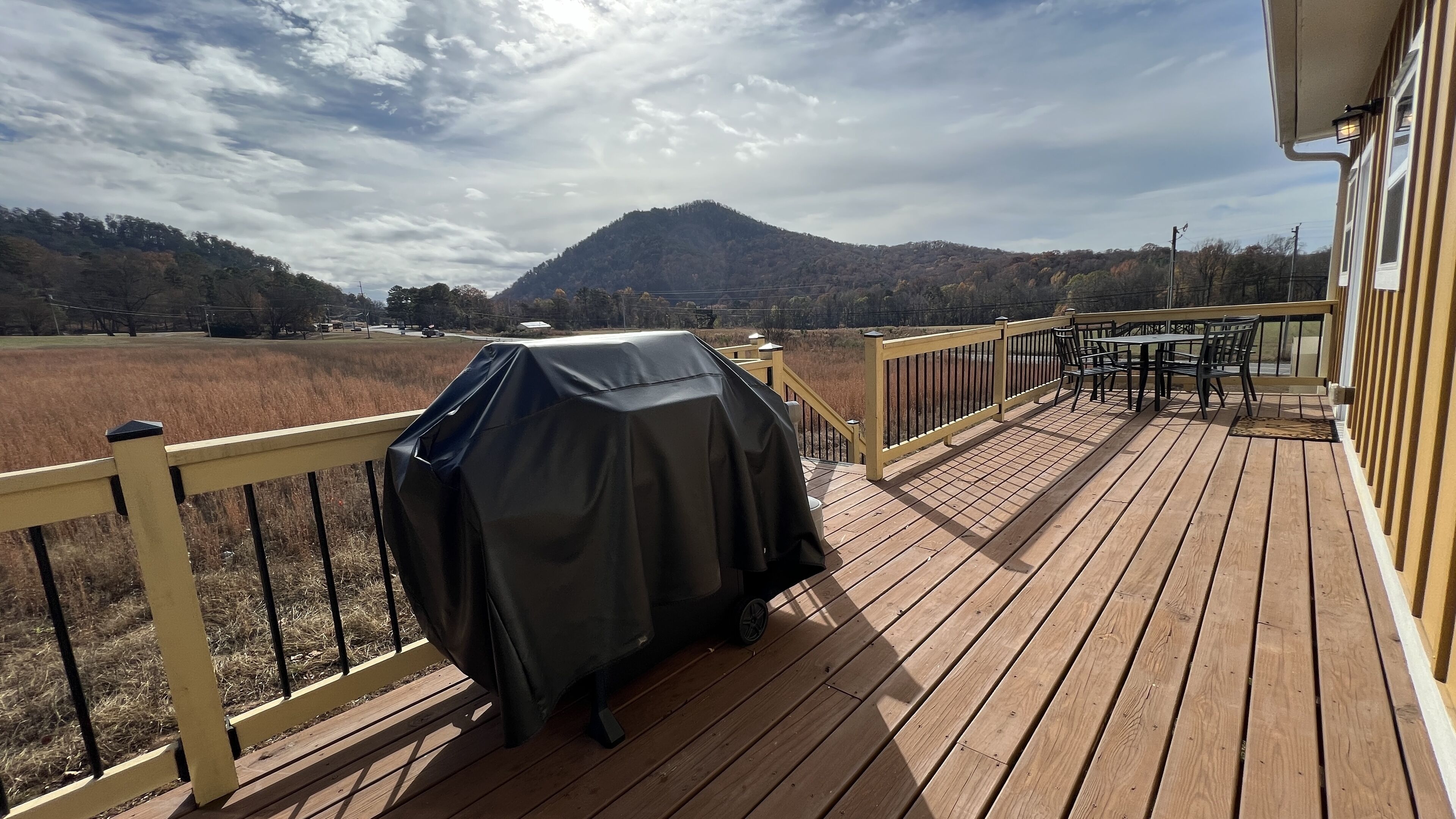 A wooden deck with a barbecue grill, outdoor furniture, and a view of a mountain and field.
