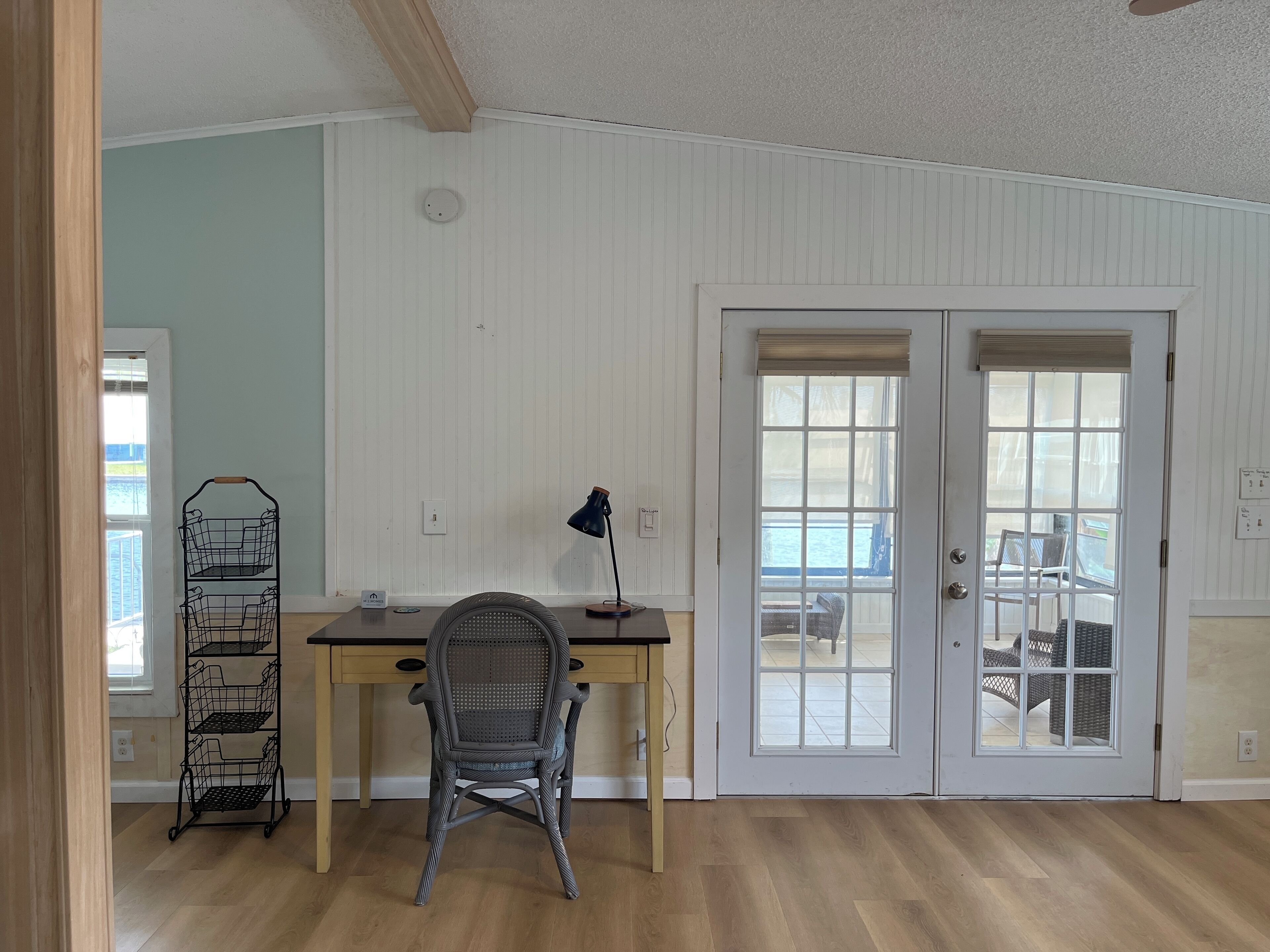 Une pièce avec un bureau en bois, une chaise en osier, une lampe de bureau noire et une étagère en fil de fer noir.