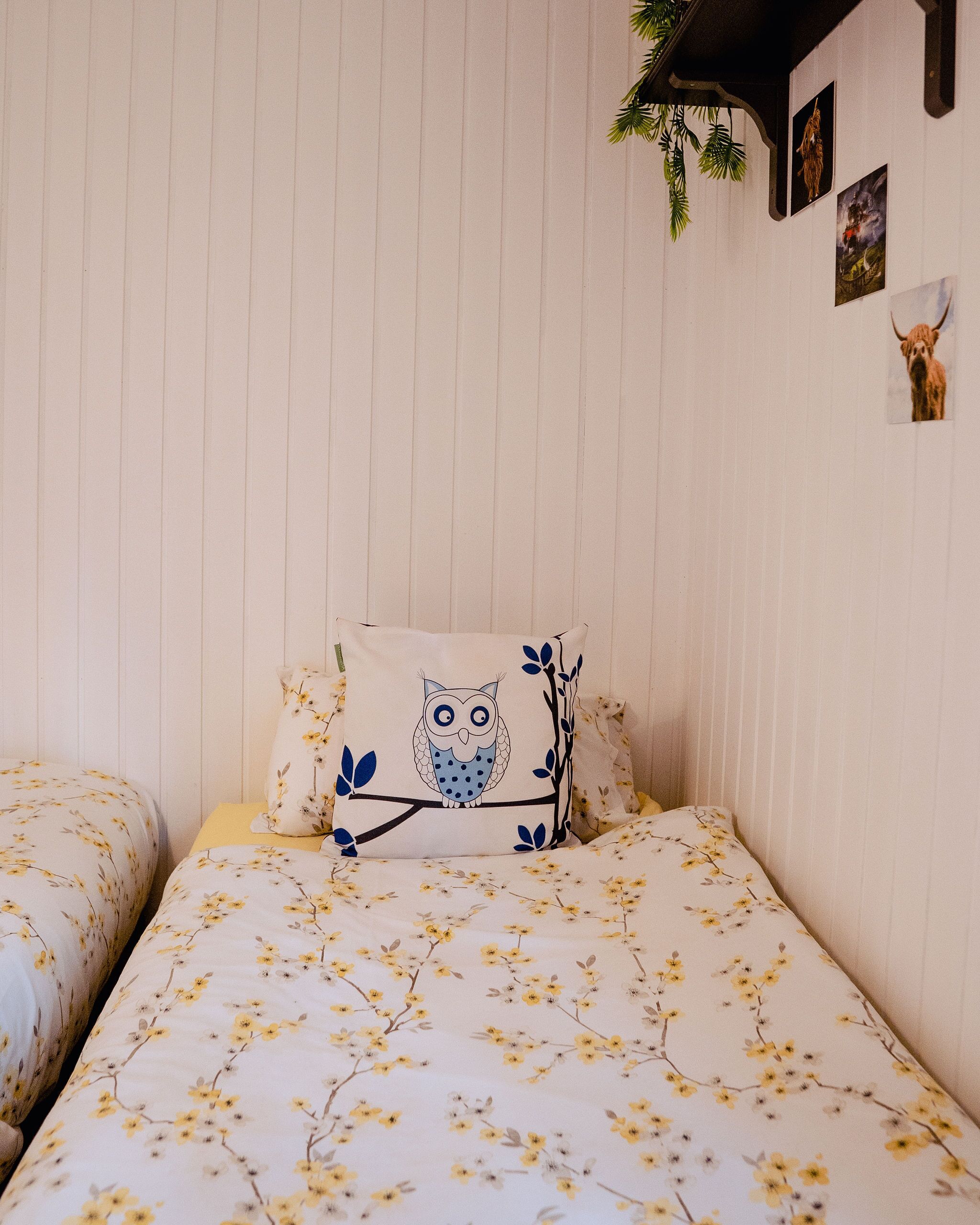 Et soveværelse med to senge, hver med blomstret sengetøj, og en pude med et ugle-motiv. Væggen har lodrette linjer og indrammede billeder.
