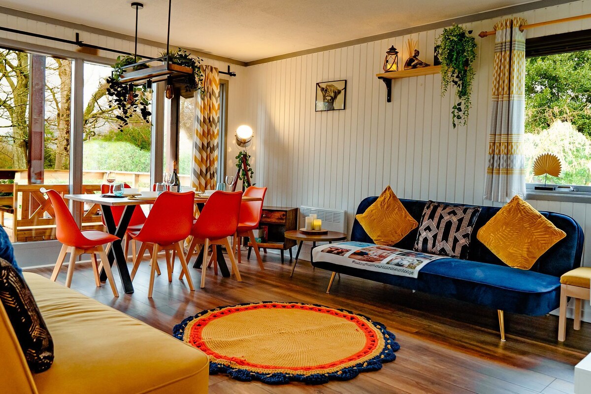 Un salón moderno con un sofá azul, sillas rojas y un sofá amarillo. Cuenta con una zona de comedor con mesa y sillas, y un ventanal con plantas y cortinas.